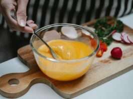 High angle of crop female beating eggs in glass bowl standing at table with ingredients for breakfast on cutting board