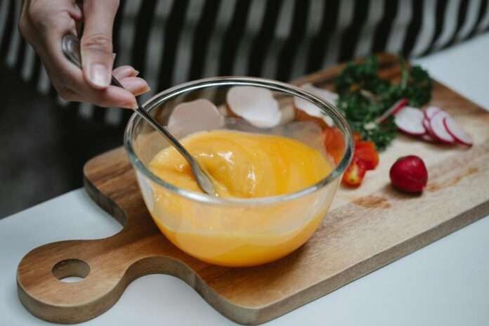 High angle of crop female beating eggs in glass bowl standing at table with ingredients for breakfast on cutting board